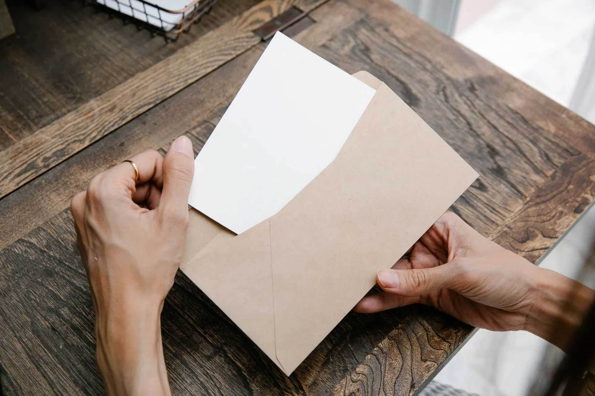 Two hands slide a blank card into a kraft envelope on a dark wooden desk.