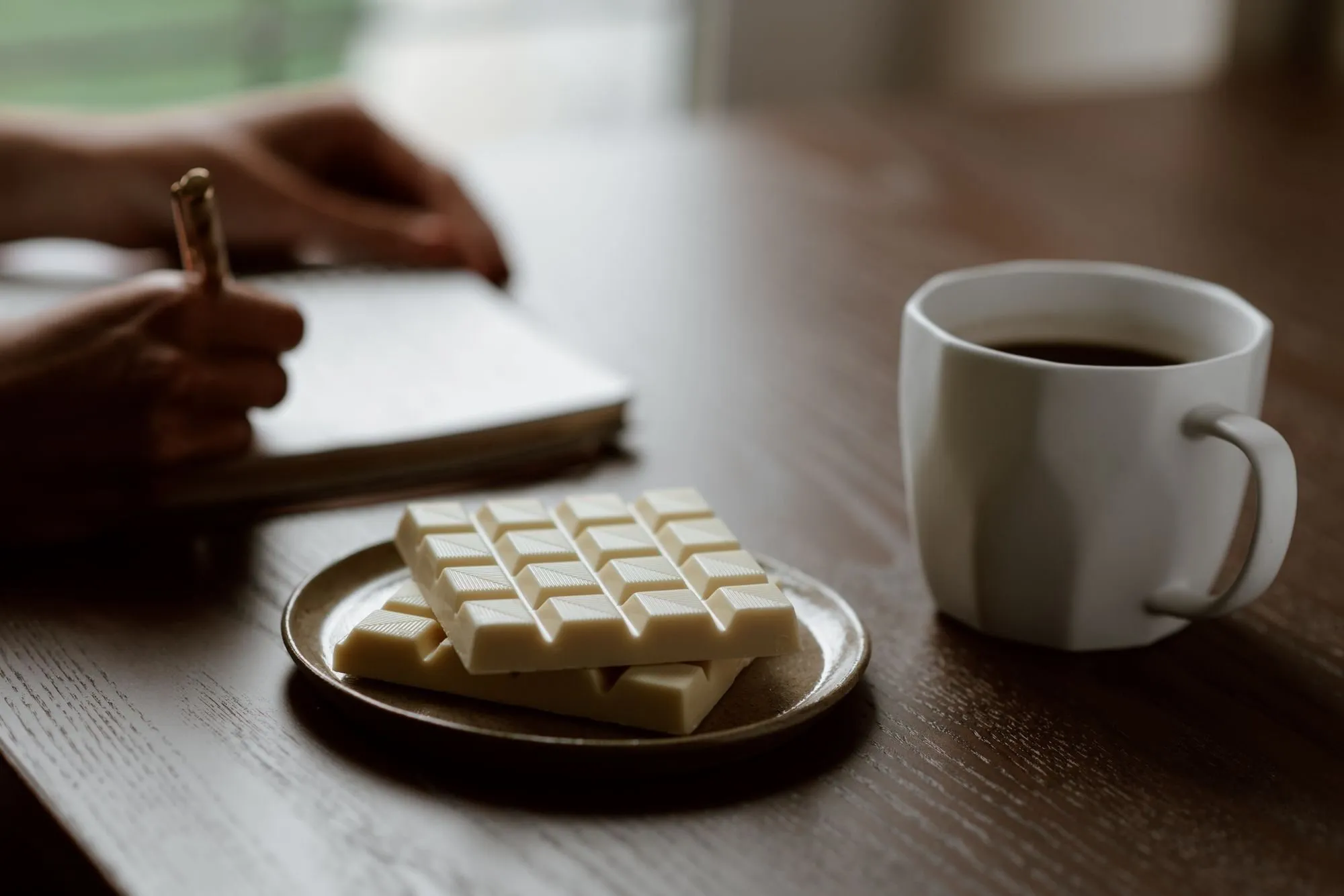 A plate of white chocolate and a cup of coffee sit beside a person writing in a notebook.
