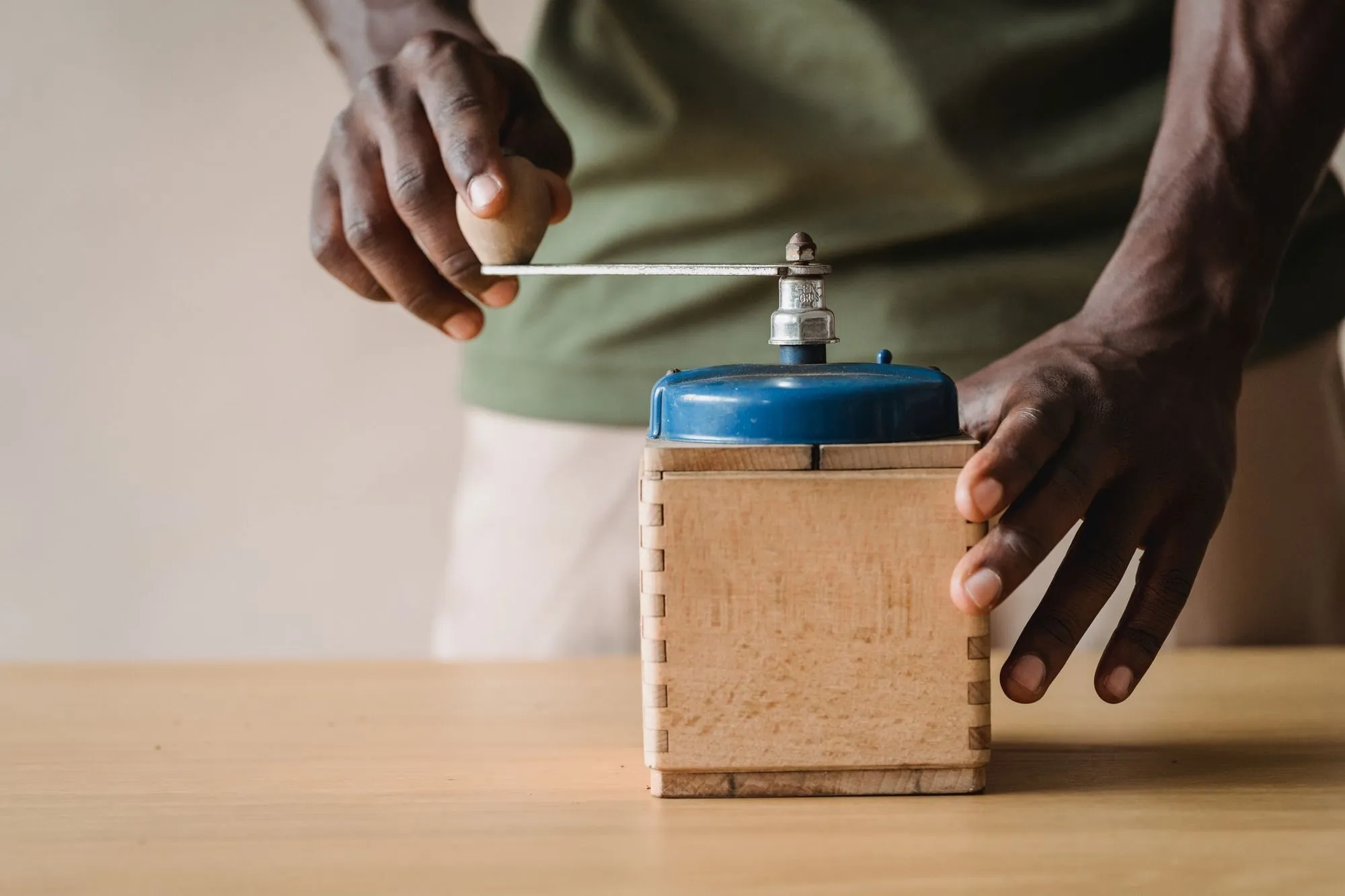 Two hands turn the crank of an old wooden coffee grinder on a table.