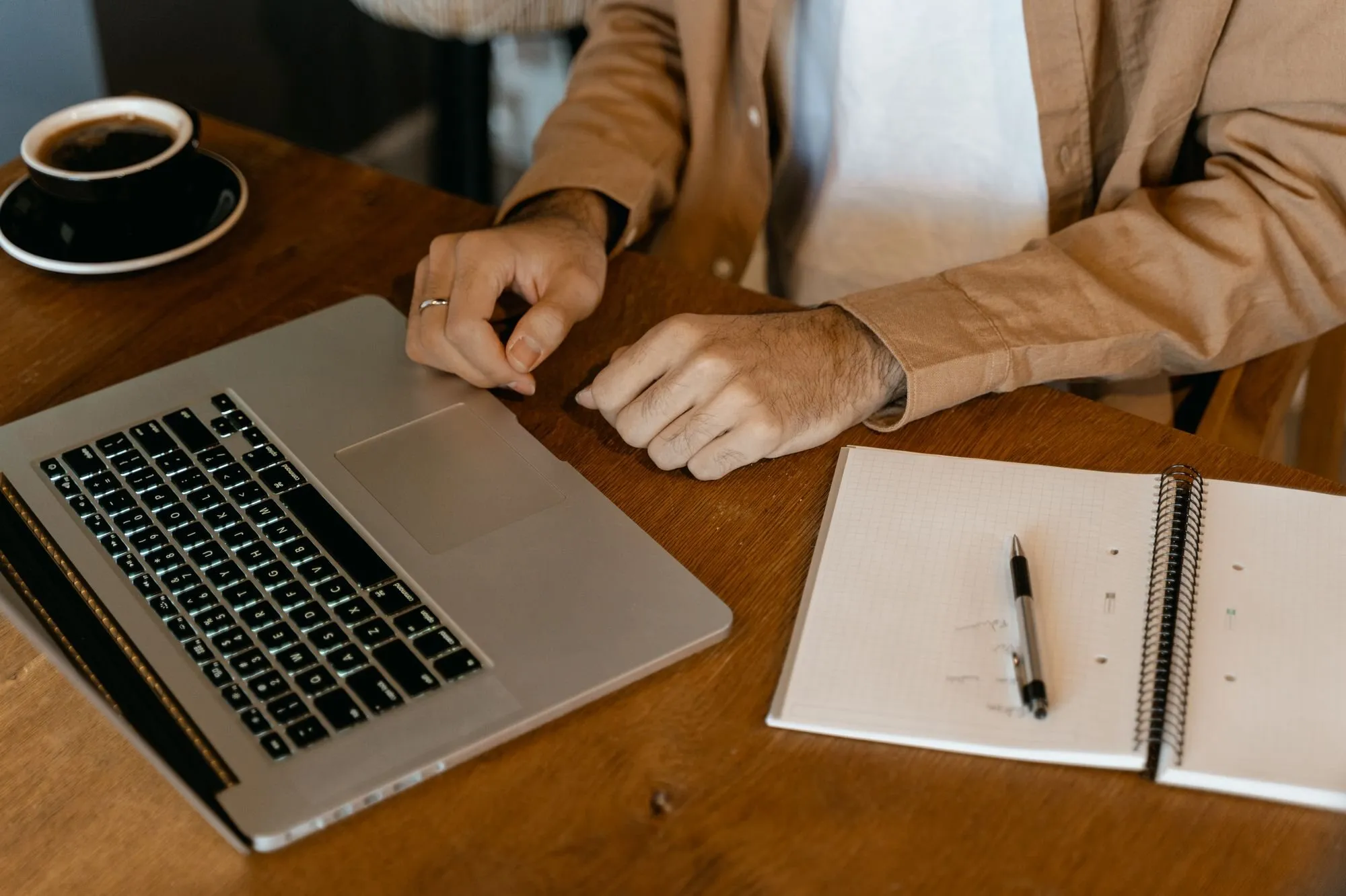 A man in a tan shirt rests his hands beside an open laptop on a wooden desk, with a notebook and a cup of coffee.