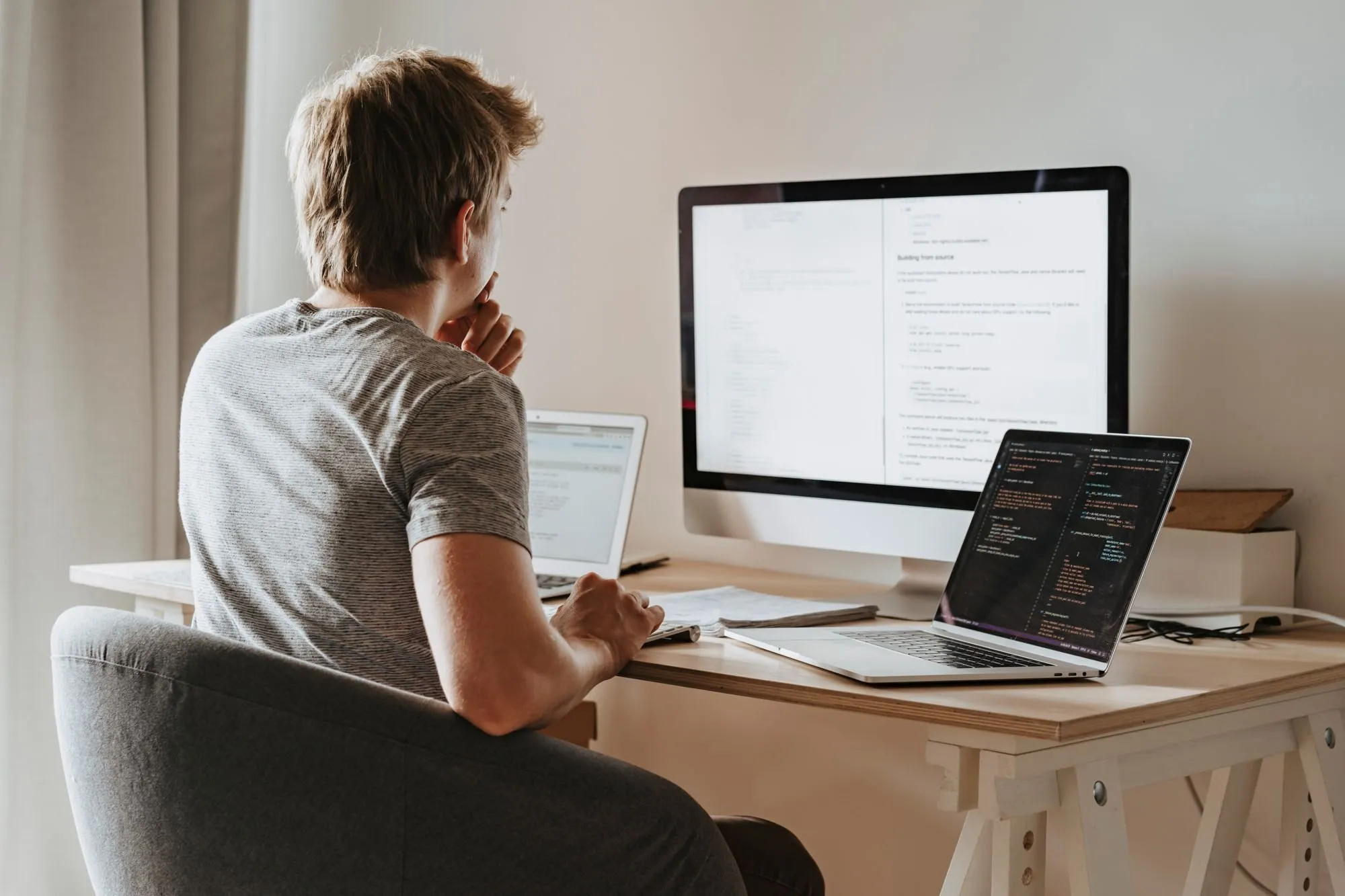 A developer sits at a light wooden desk with a large monitor and two laptops, thinking with his hand at his chin.