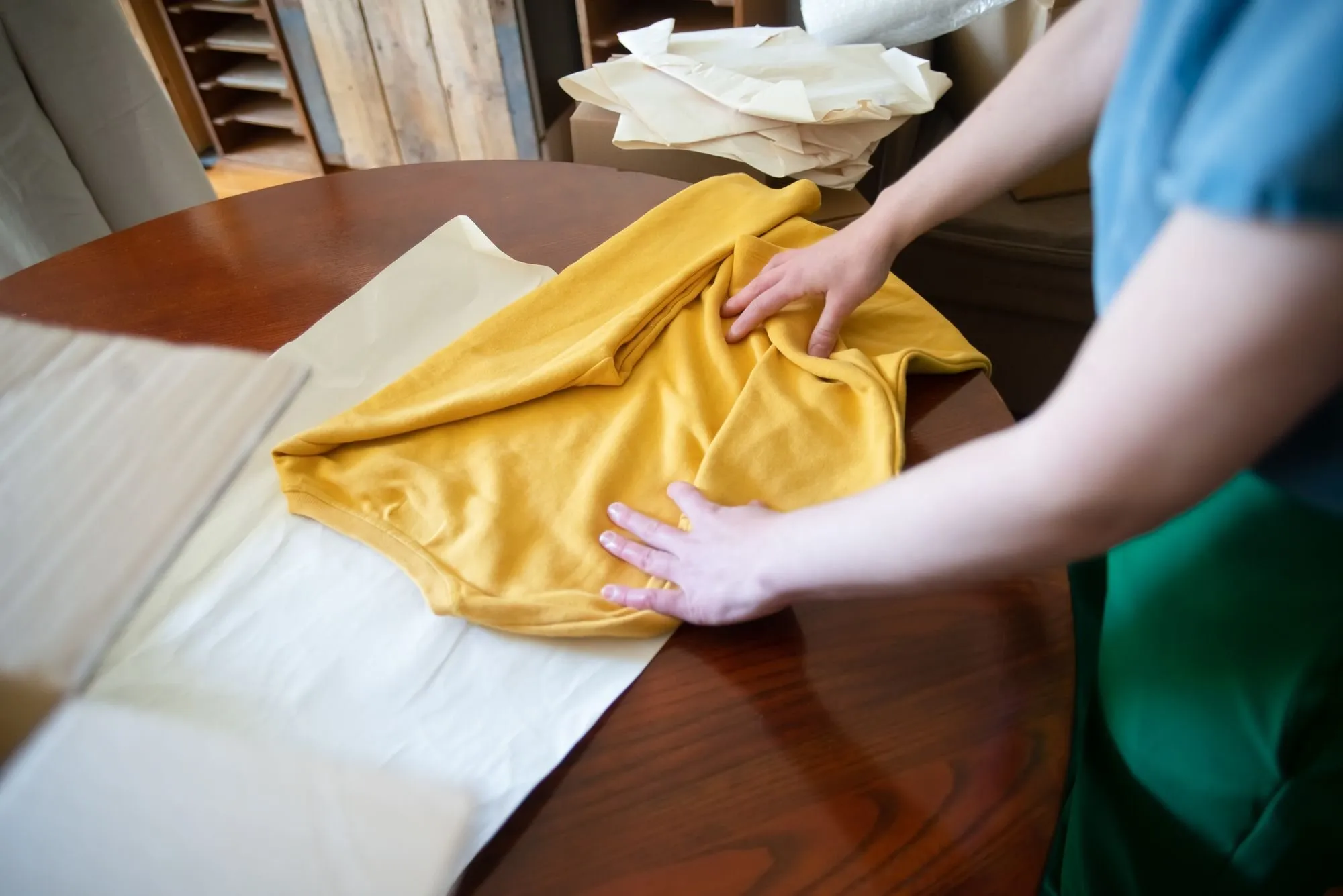 Hands fold a yellow shirt onto tissue paper on a round wooden table.