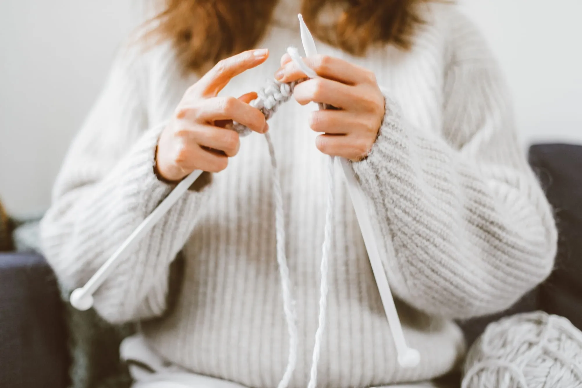 Two hands knit pale yarn on long needles against a cream sweater.