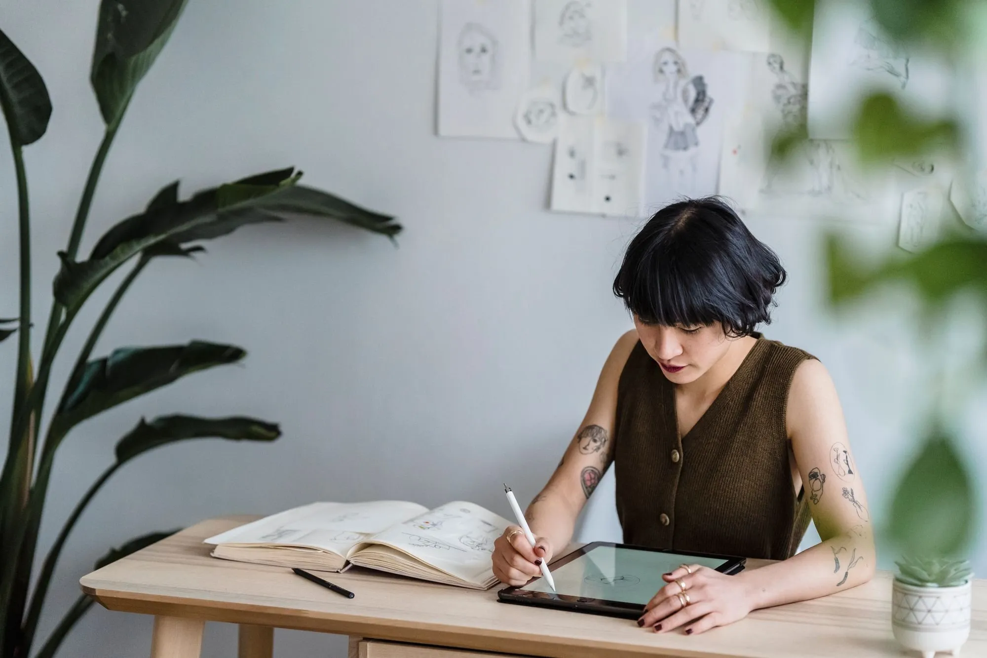 An illustrator draws on a tablet at a wooden table, with sketches pinned to the wall and a plant beside her.