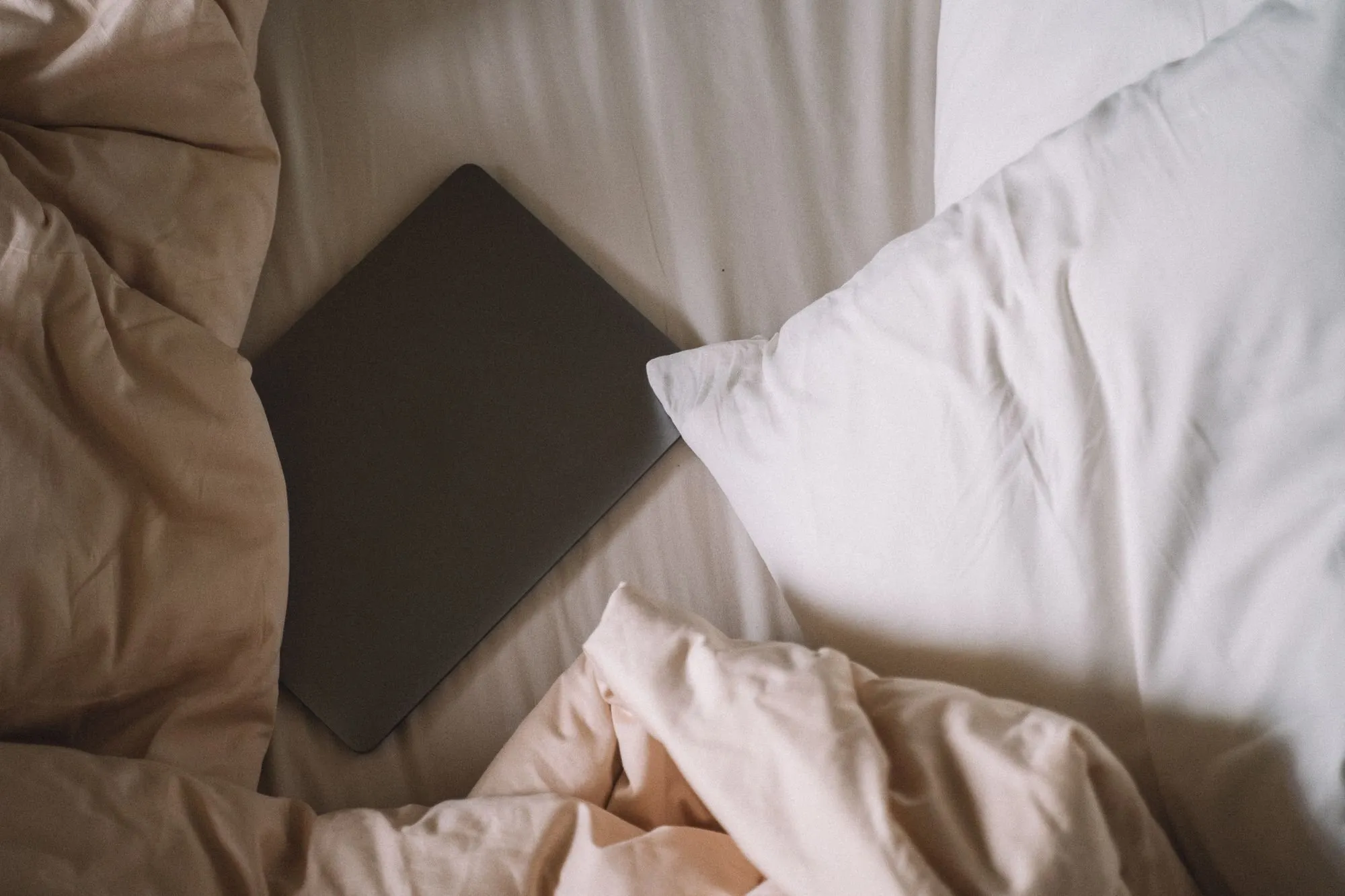 A closed laptop rests on an unmade bed between soft pillows.