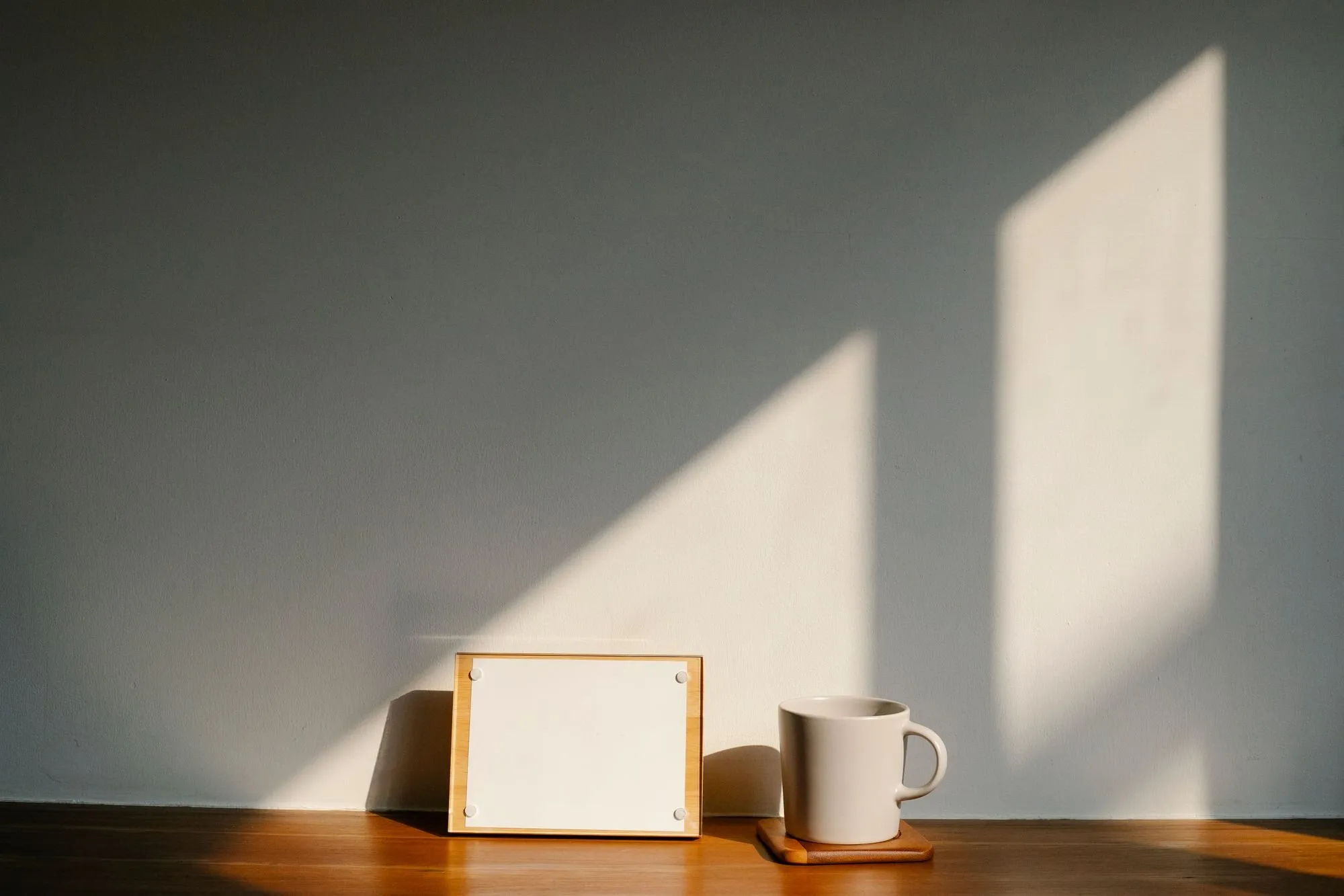 A white mug and a small blank frame stand on a wooden shelf, with window light falling across the wall.
