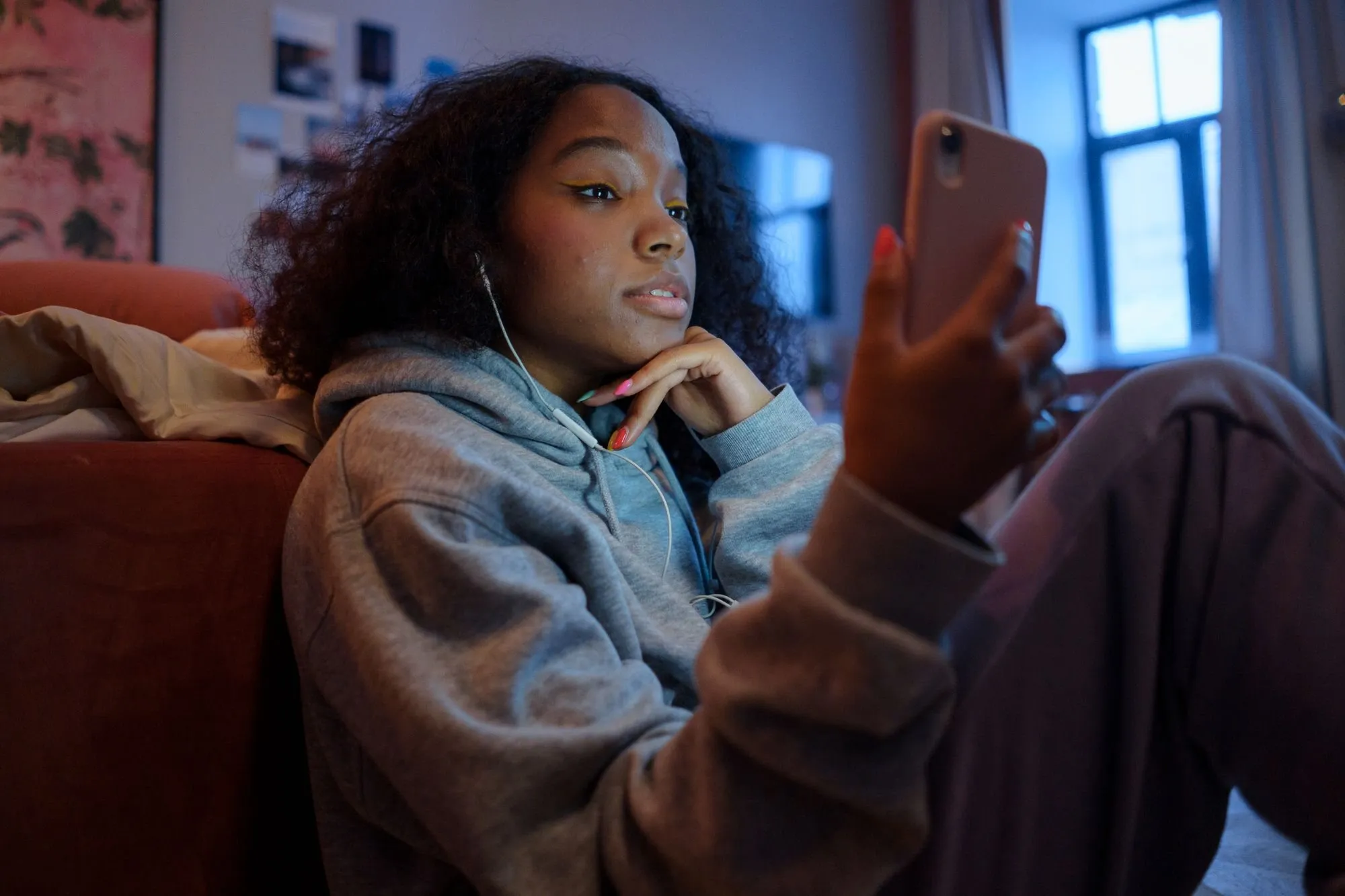 A young woman in a hoodie sits against a sofa with earphones in, looking at her phone.