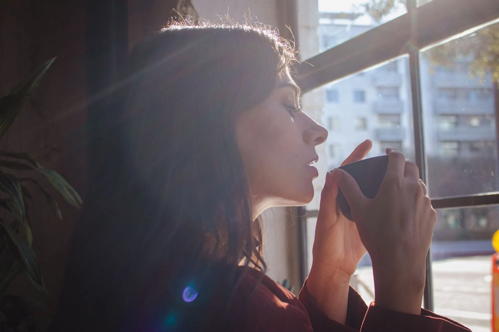 A woman sips from a mug with her eyes closed beside a sunlit window.
