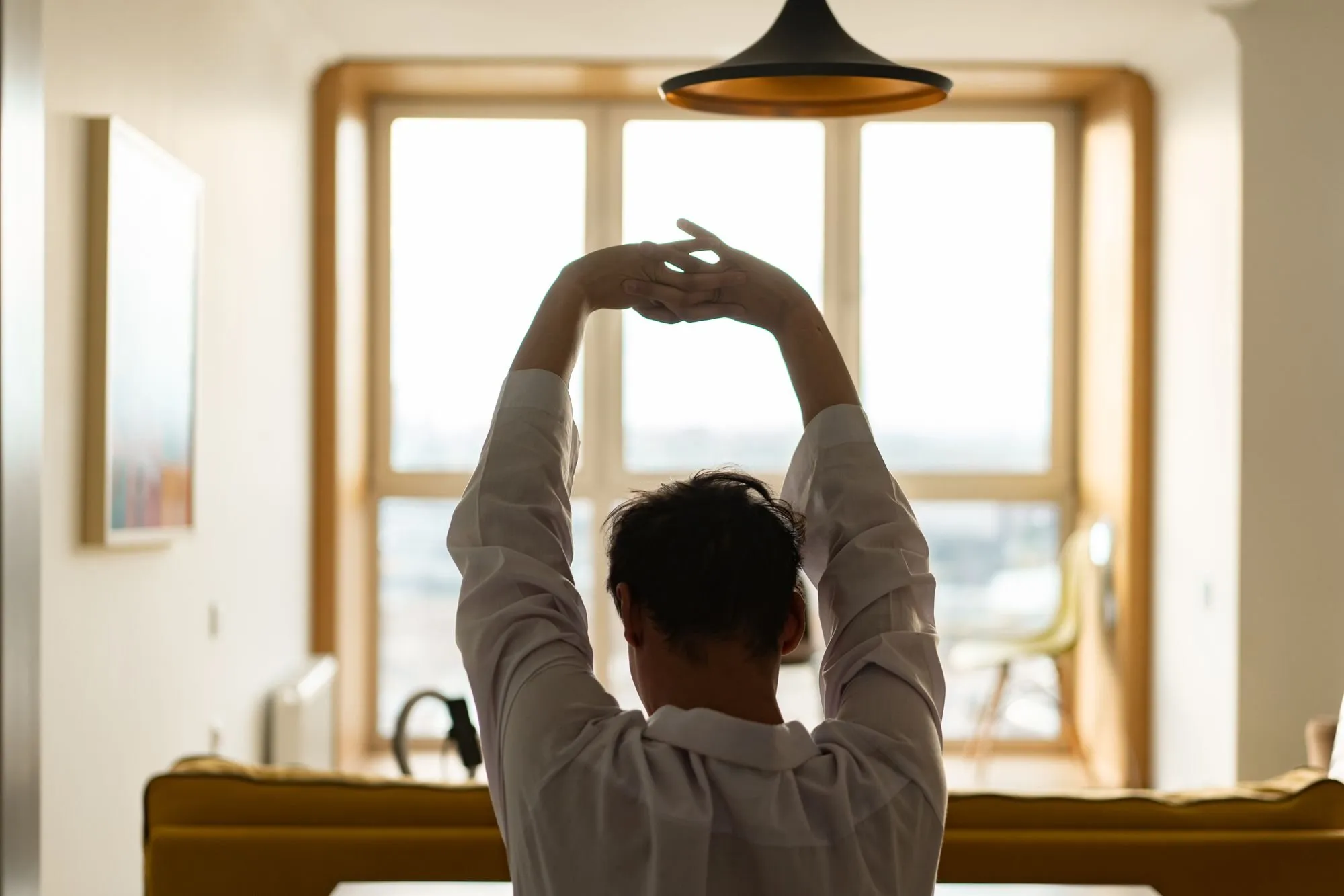 A man stretches his arms above his head in front of a bright window at home.